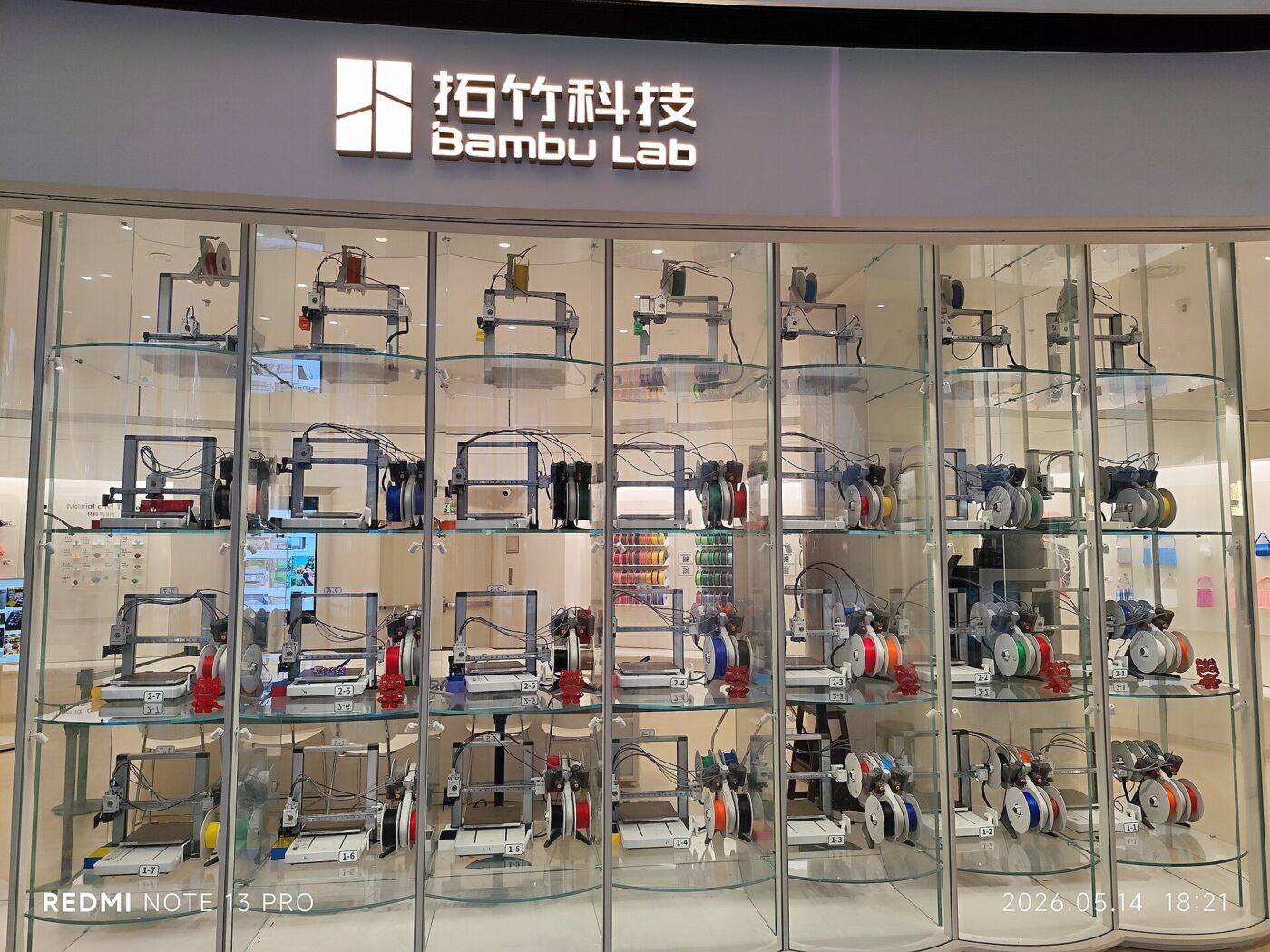 Glass display shelves at a Bambu Lab store hold rows of desktop 3D printers and filament spools