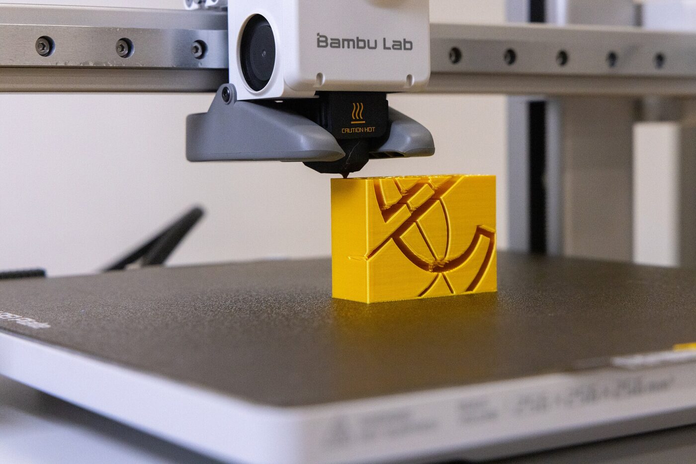 A Bambu Lab 3D printer's toolhead prints a yellow block on a textured build plate