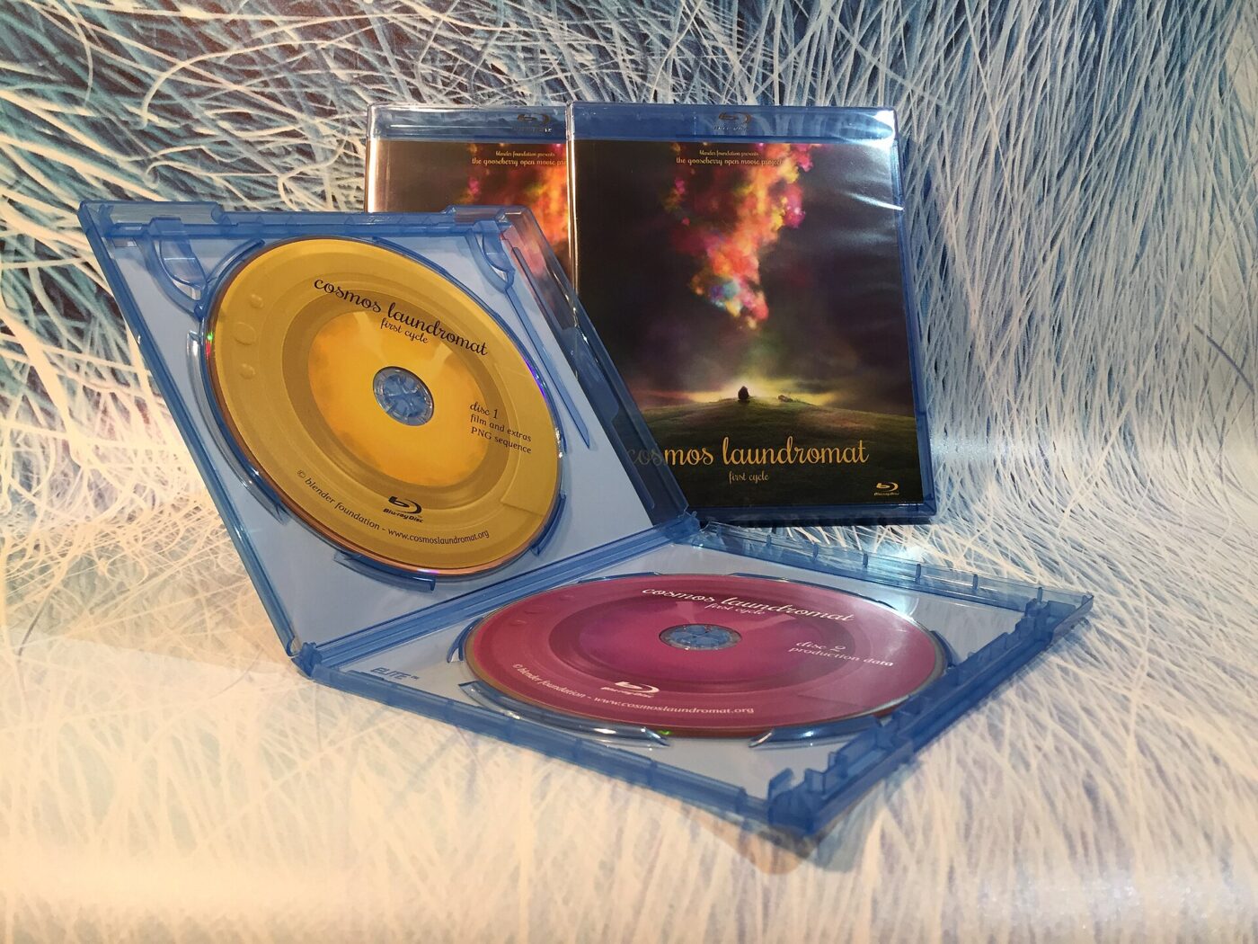 An open Blu-ray case holding two printed discs, with boxed copies of the same film behind it