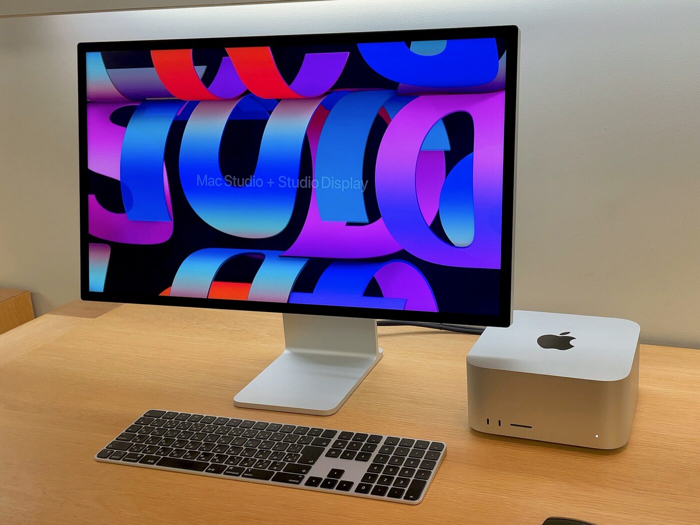 An Apple Studio Display on a wooden table next to a Mac Studio and a keyboard