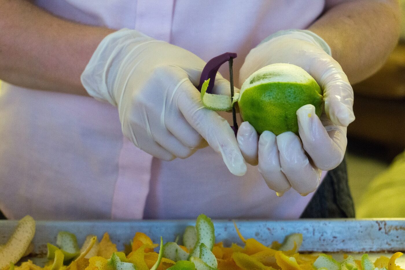 Gloved hands run a Y-shaped peeler down a lime over a tray of peel.