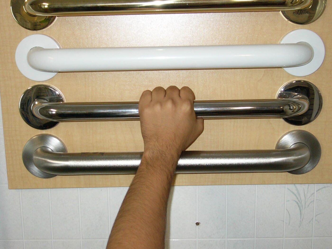 A hand grips one of four grab bars in different finishes mounted on a display board