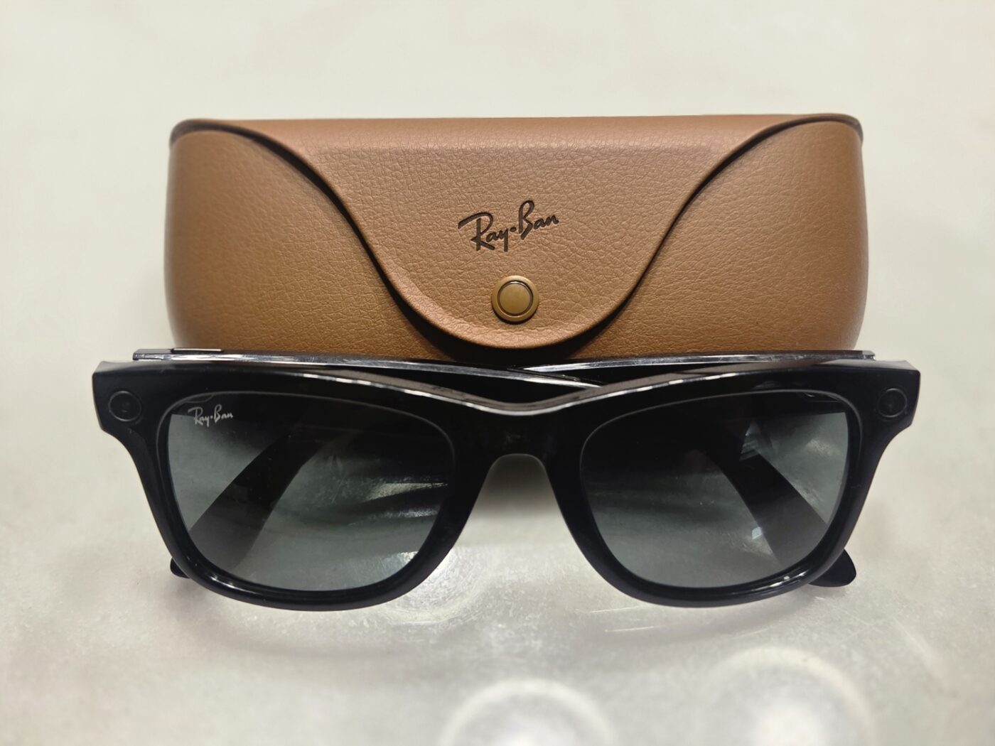 Sunglasses-style smart glasses with a built-in camera at the frame corner, shown in front of a tan case