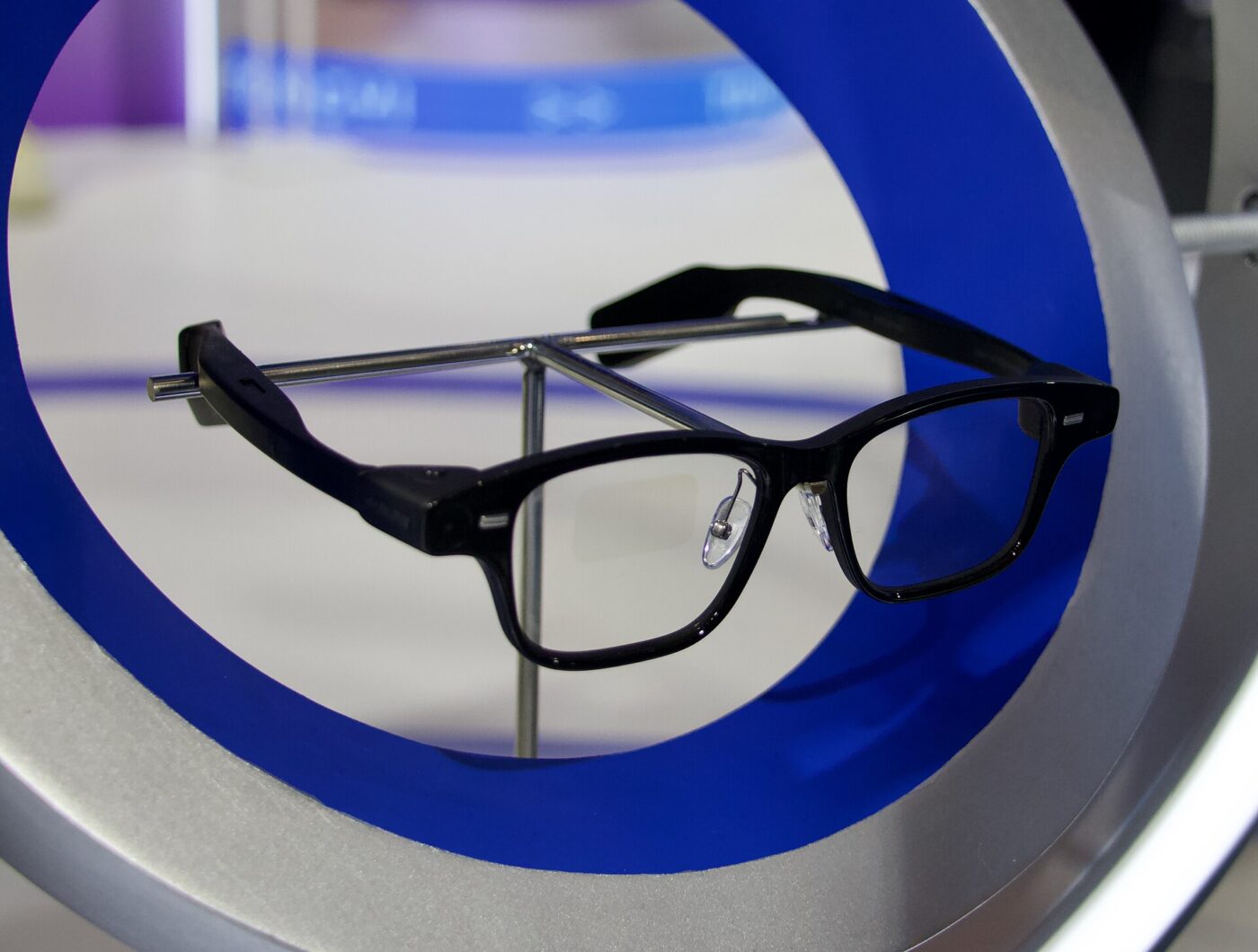 Black smart glasses with thick temple arms resting on a metal stand inside a blue ring display