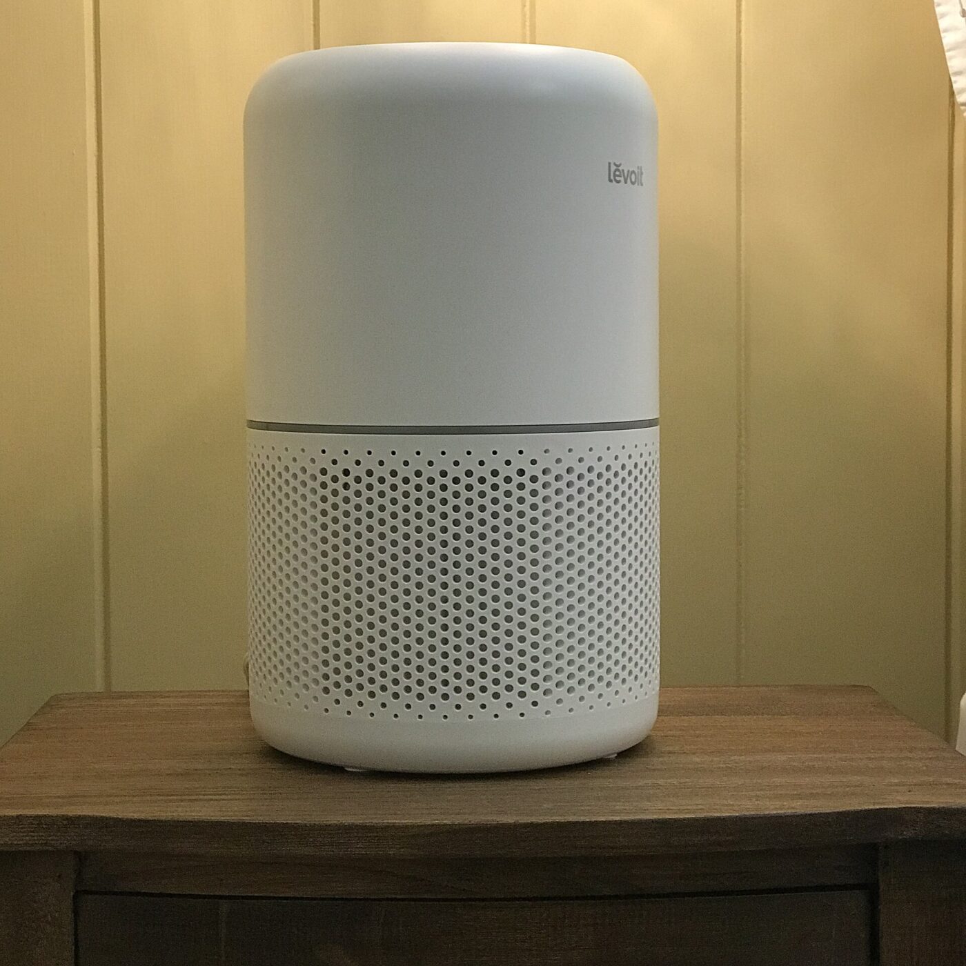 A white cylindrical room air purifier on a wooden nightstand against a paneled wall