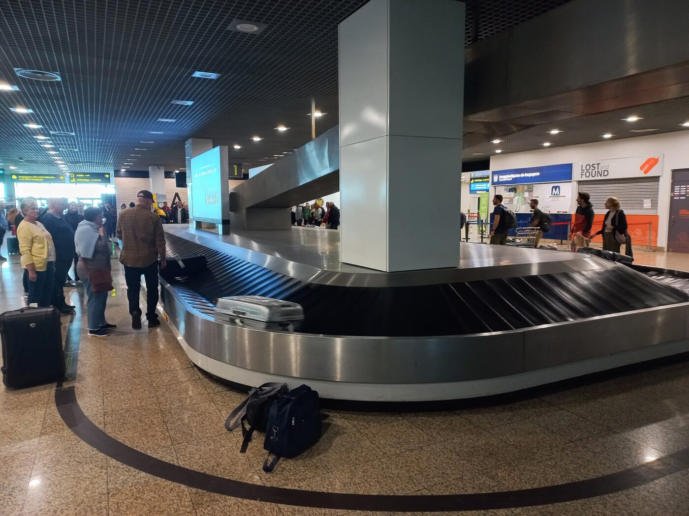 Travelers wait beside an airport baggage carousel as checked suitcases come off the belt