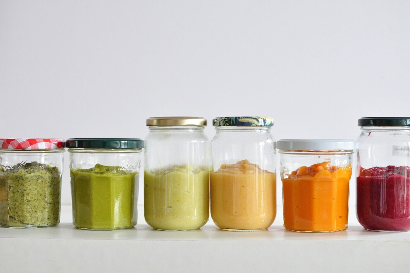 Glass jars of smooth vegetable and fruit purees lined up in a row on a white shelf