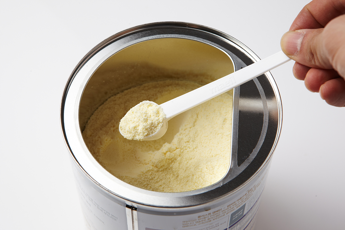 Scoop lifting powdered infant formula from an open metal can against a white background