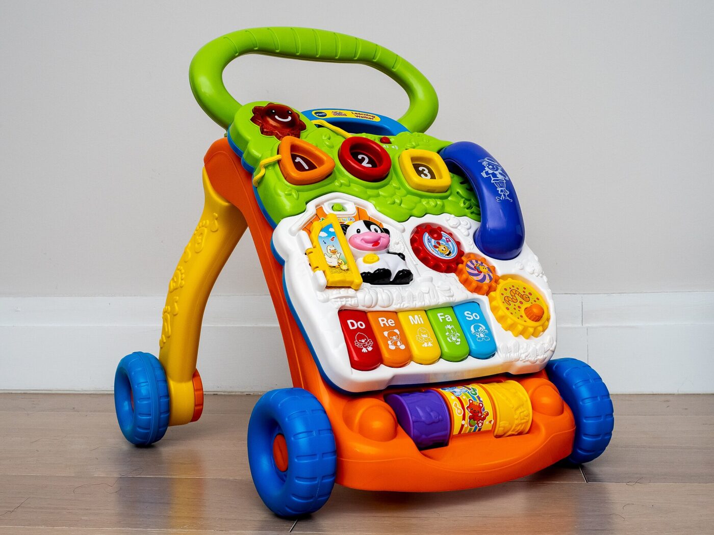 A brightly colored plastic push walker with a toy panel, handle and four wheels.
