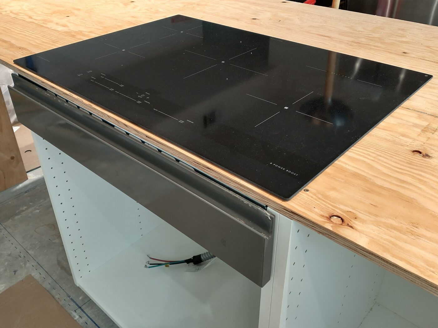 A black glass induction cooktop set into a plywood countertop during installation, with wiring visible below