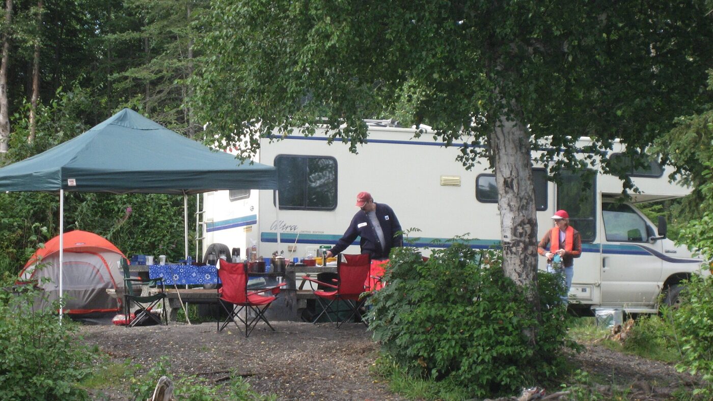 A wooded campsite with a trailer, canopy, tent and folding chairs around a table