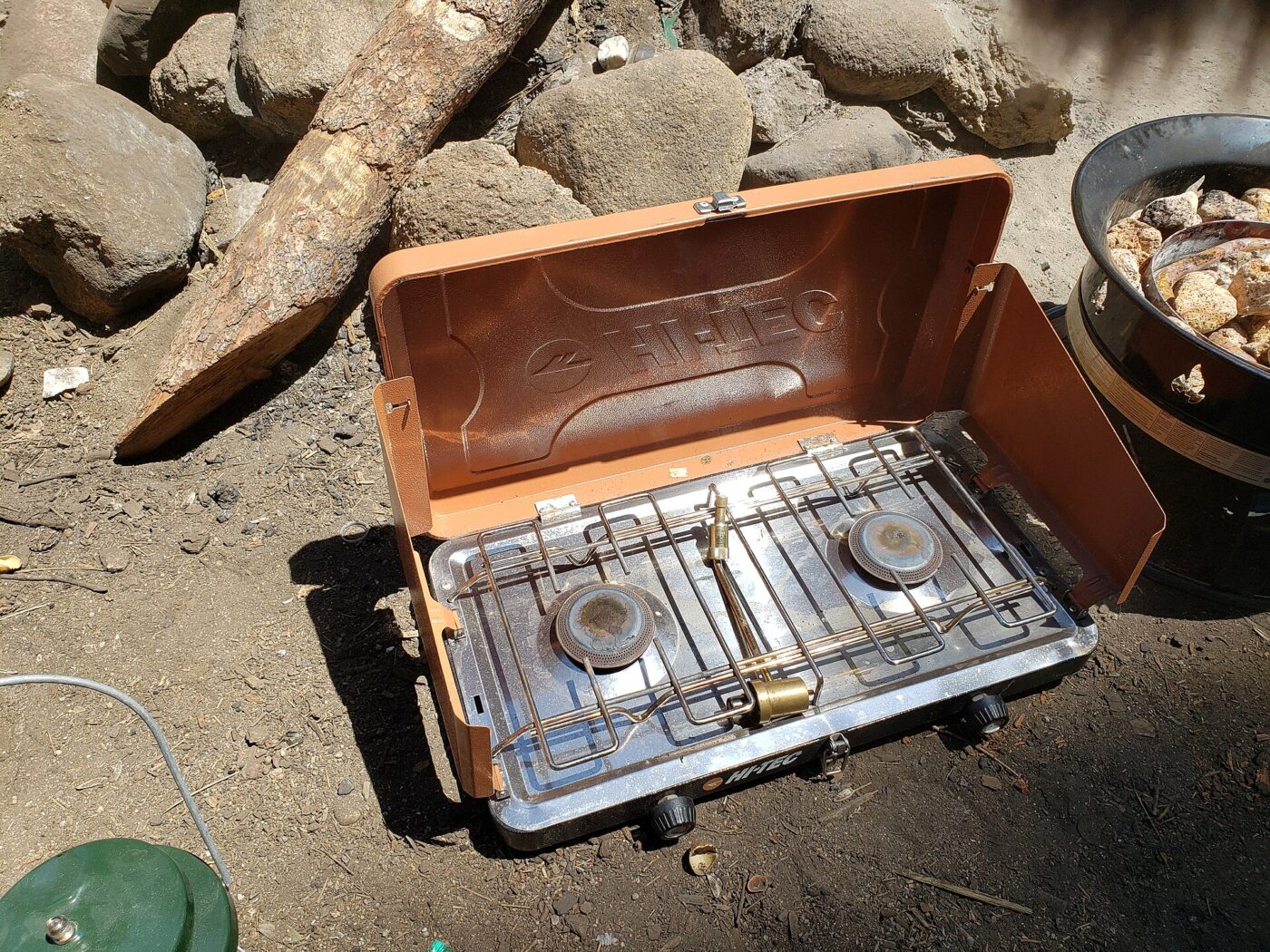 A two-burner camp stove fueled by a bottled gas cylinder, set up on bare ground
