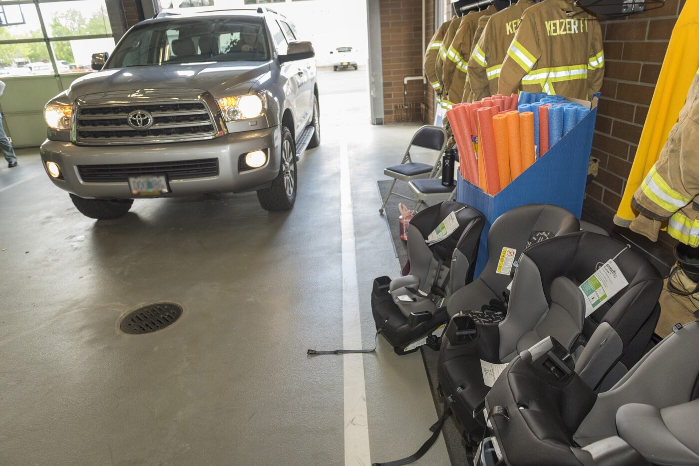 New child car seats with hang tags lined up in a fire station bay as an SUV pulls in