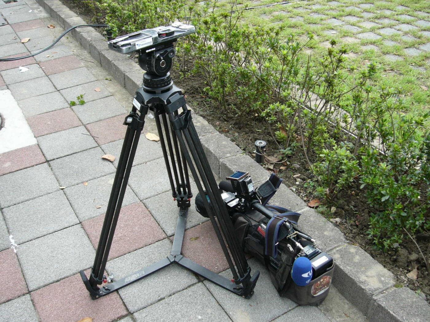 A video tripod with a fluid head and leg spreader stands on a sidewalk beside a camera.