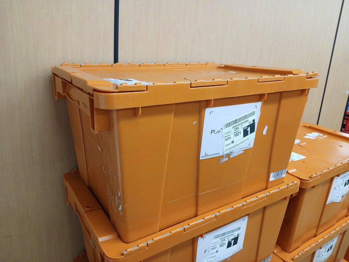 Stackable orange plastic storage crates with hinged lids, piled against a wall.