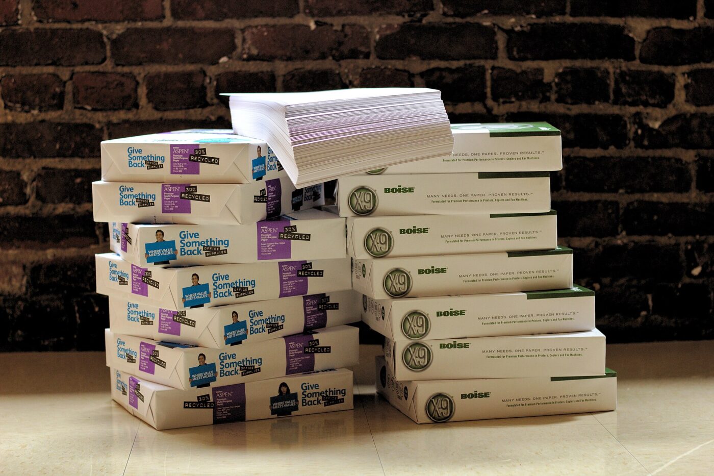 Fifteen reams of copy paper stacked in short piles on the floor against a brick wall