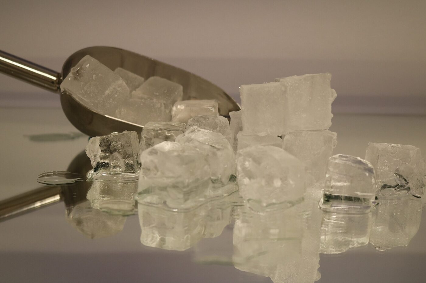 Clear ice cubes piled next to a metal scoop on a reflective surface.