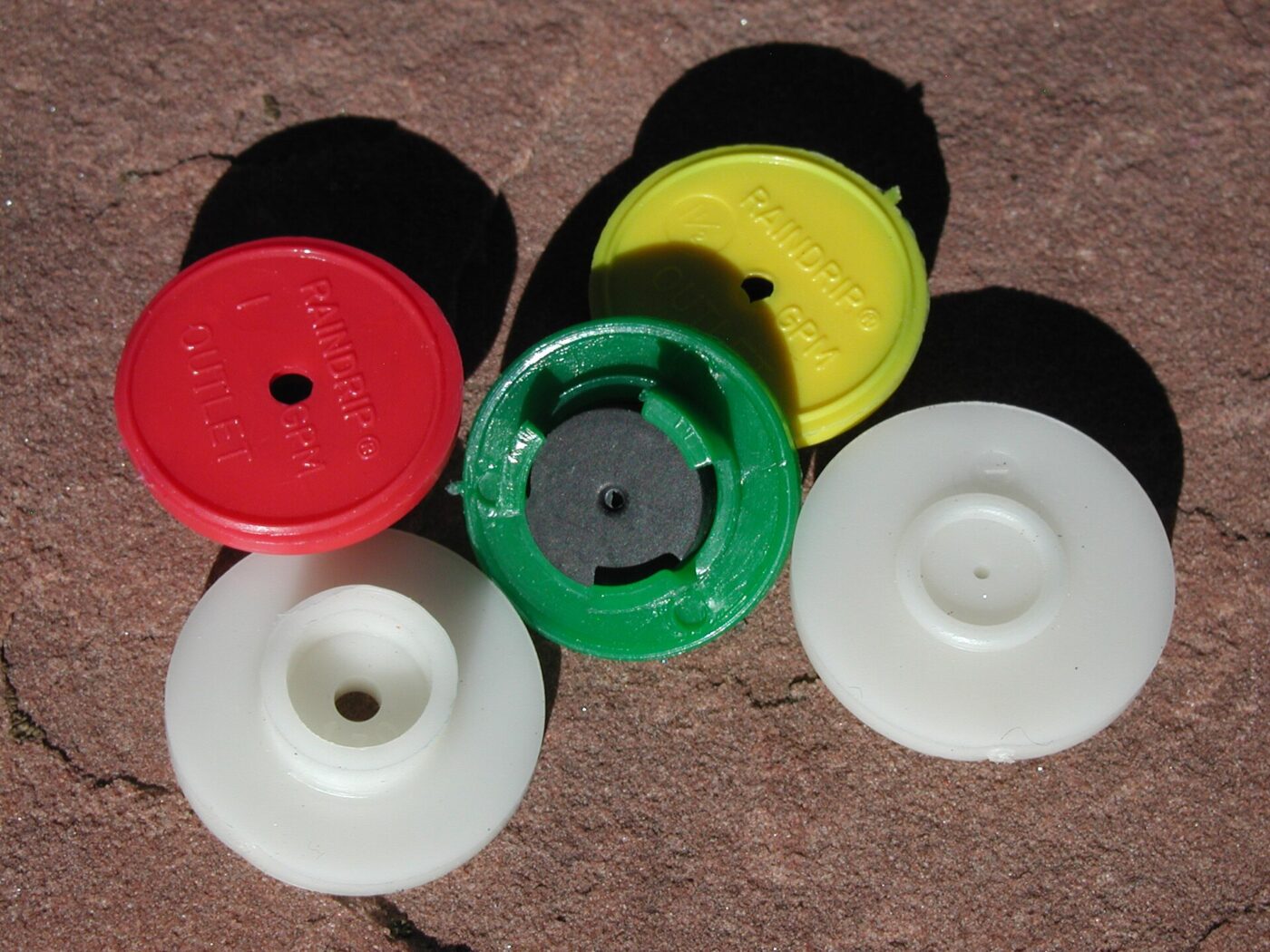 Five plastic drip irrigation emitters in red, yellow, green and white on stone.