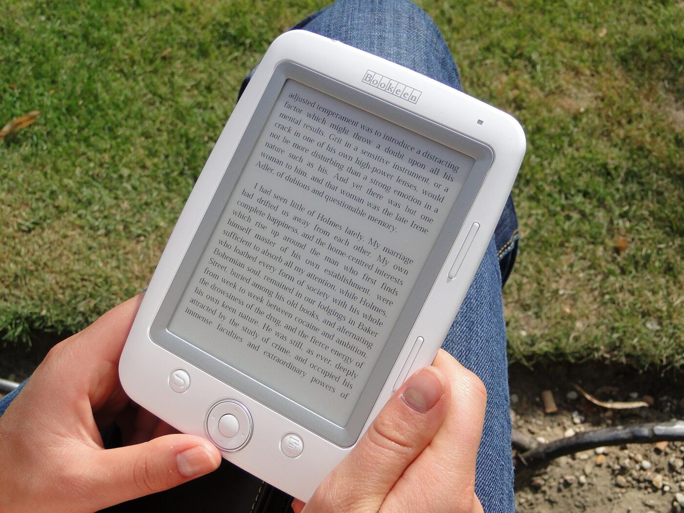 A handheld e-paper reader showing a page of text, held outdoors above a lawn