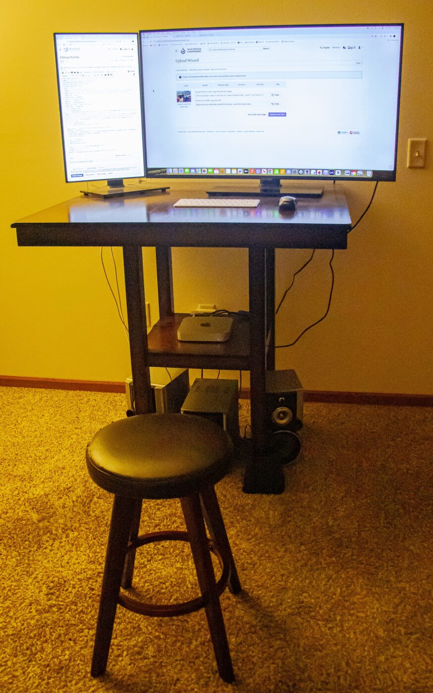Two monitors on a raised standing desk in a home office, with a tall stool alongside.