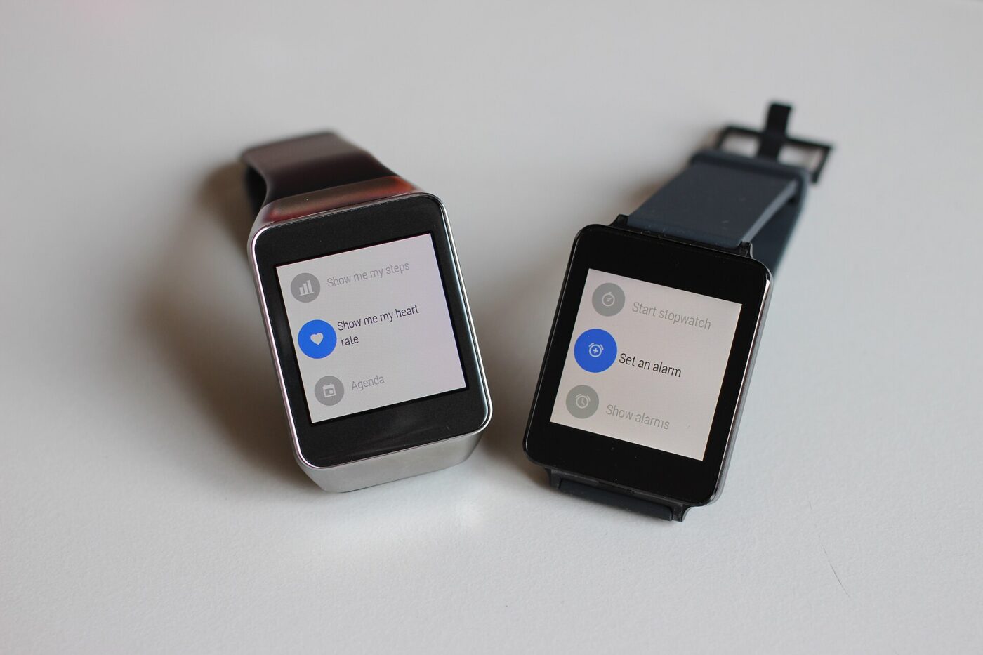 Two square-faced smartwatches showing on-screen menus of built-in features such as alarms and heart rate