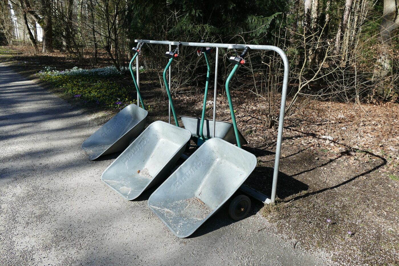 Three galvanized steel wheelbarrows with green handles lined up on a paved path.