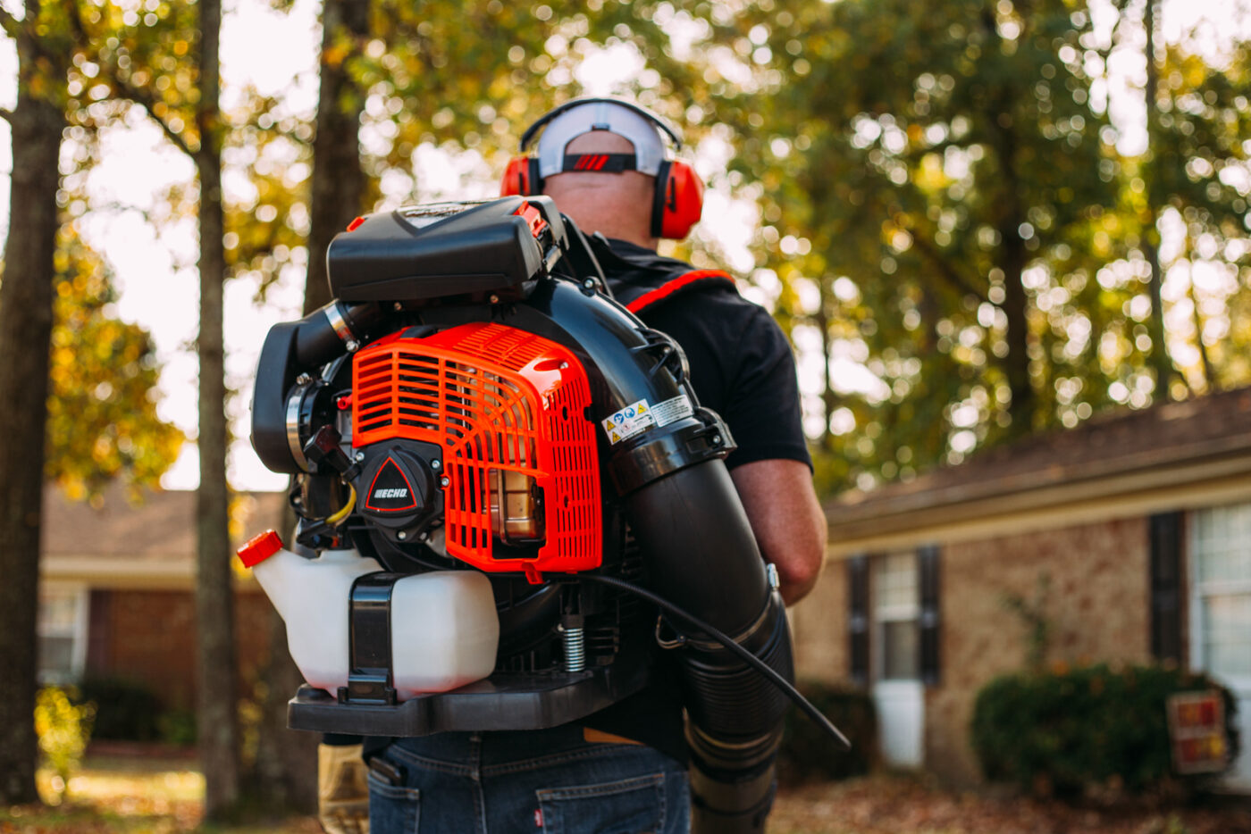 A worker wearing ear protection carries a gas-powered backpack leaf blower past a brick house