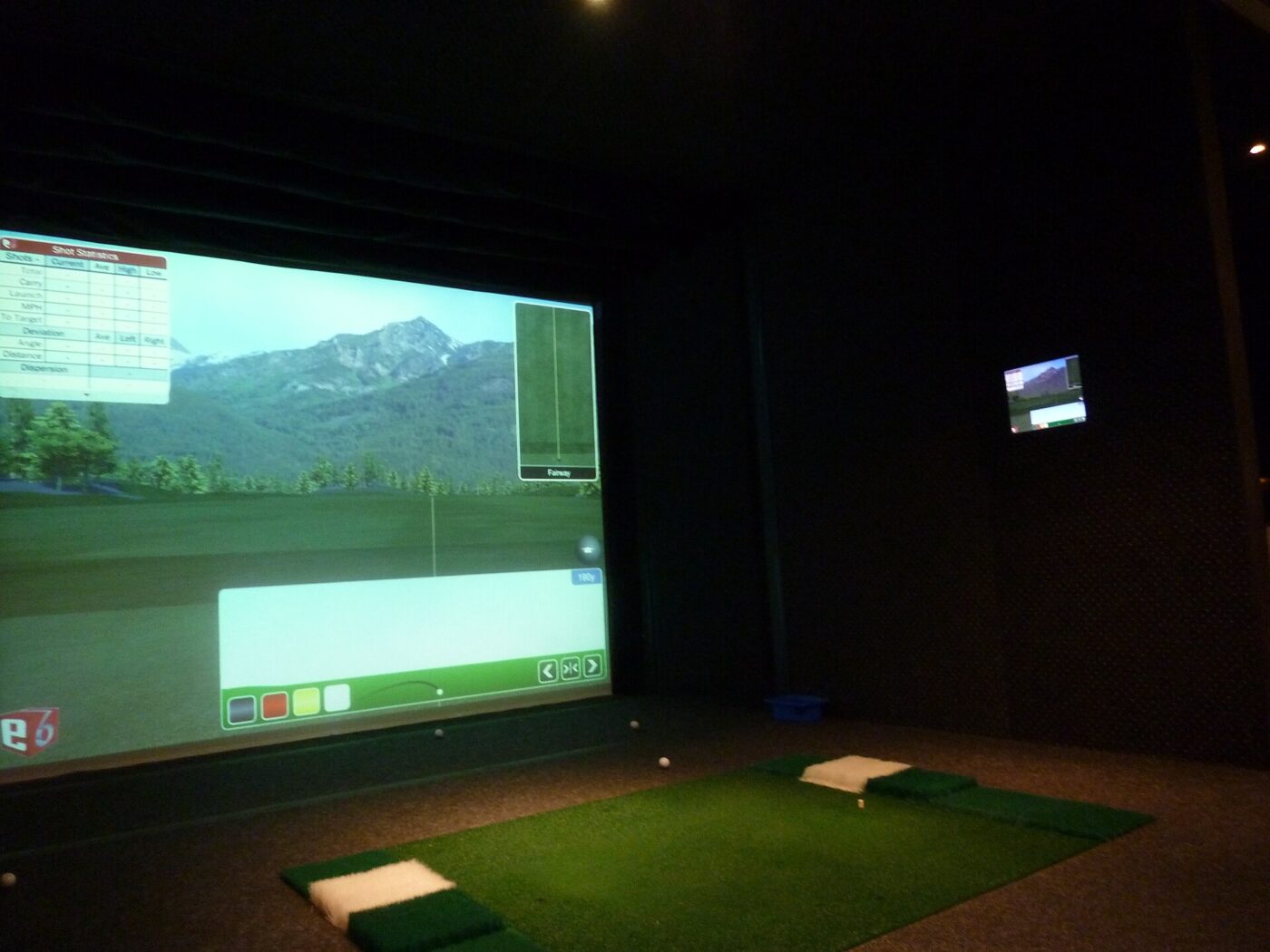 A darkened golf simulator room with a virtual course projected on the screen and turf hitting mats