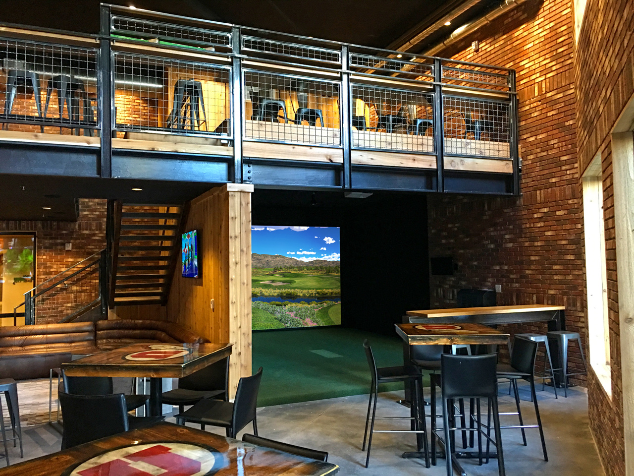 A golf simulator bay with a projected fairway on screen inside a brick-walled lounge with tables