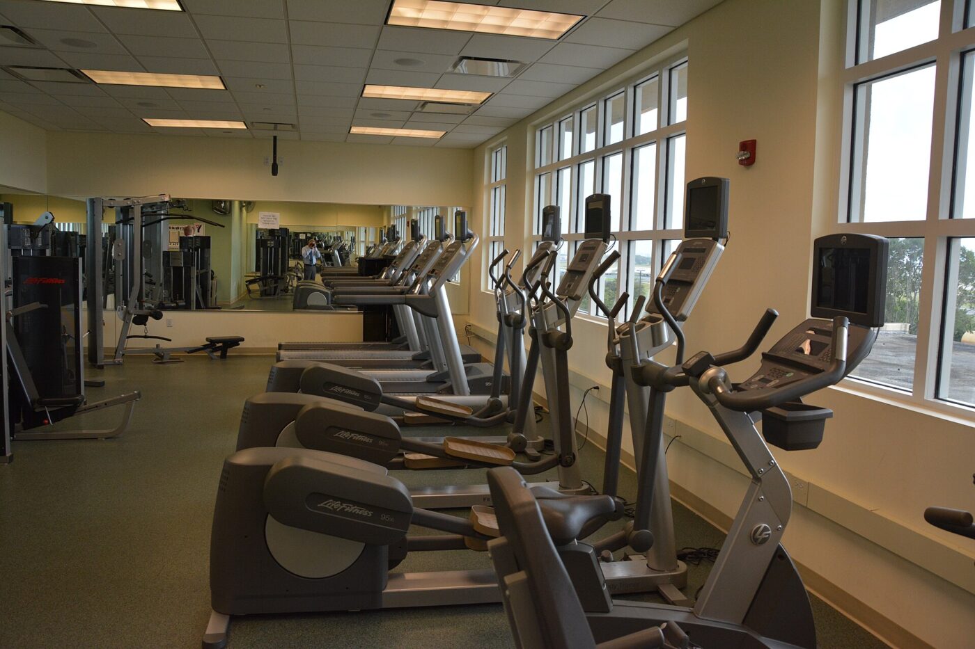 Cardio machines and weight equipment lined up beside tall windows on a gym floor.
