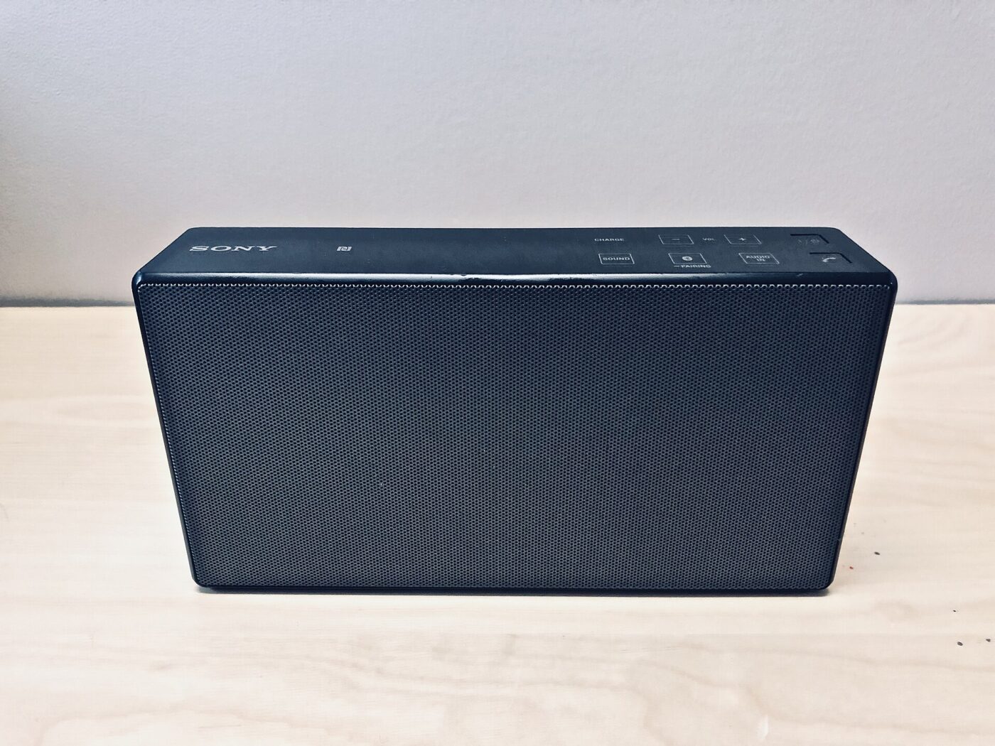 A black rectangular portable speaker with a fabric grille sits on a pale wooden surface.