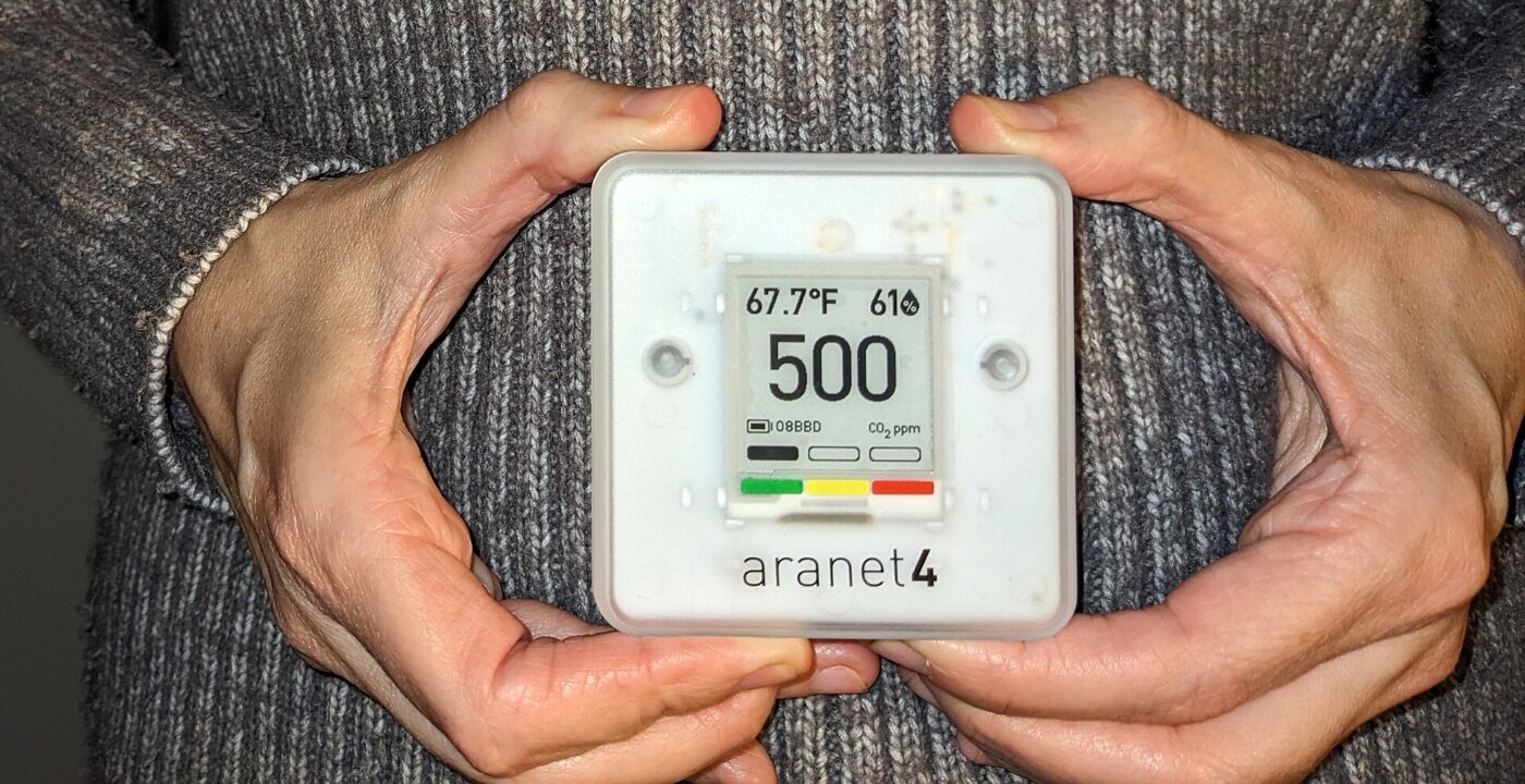 Two hands holding a small indoor air quality monitor whose screen shows a numeric reading