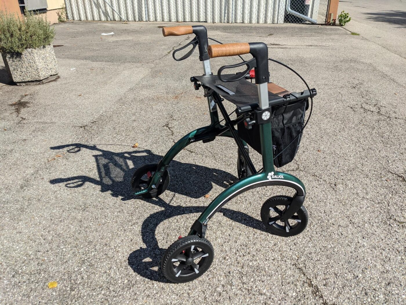 A green four-wheeled rollator walker with a seat, storage bag and wooden handles parked on pavement