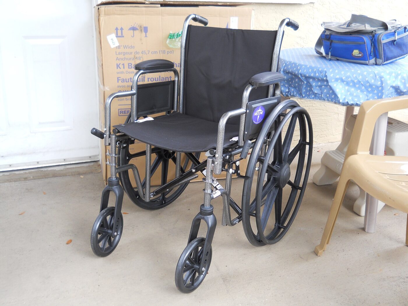 A folding manual wheelchair with padded armrests stands on a patio beside its shipping carton.