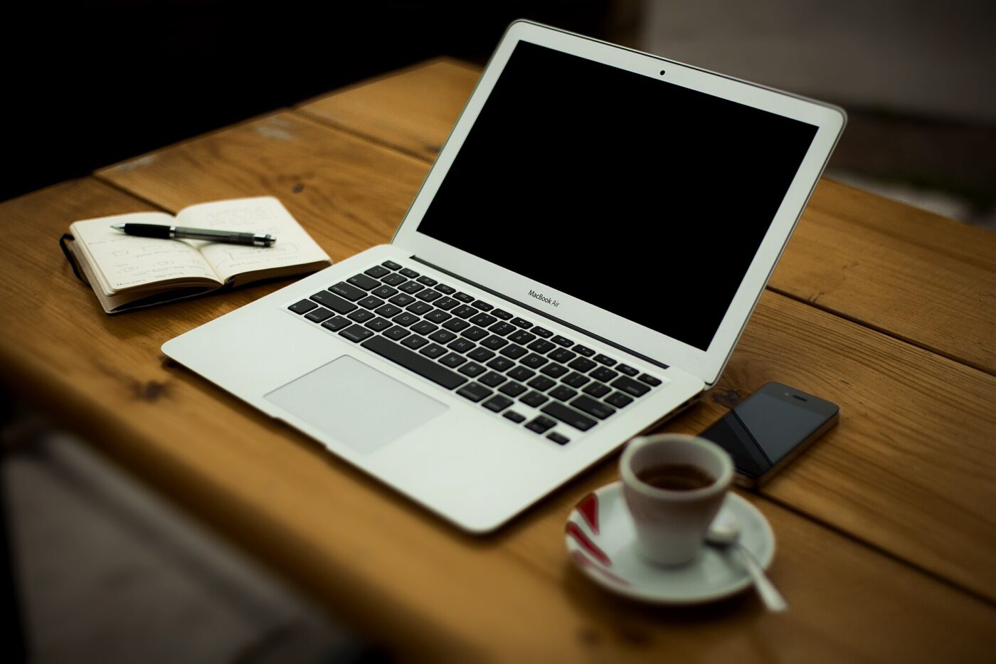 An open laptop on a wooden desk beside a notebook, pen, smartphone and a small cup of coffee