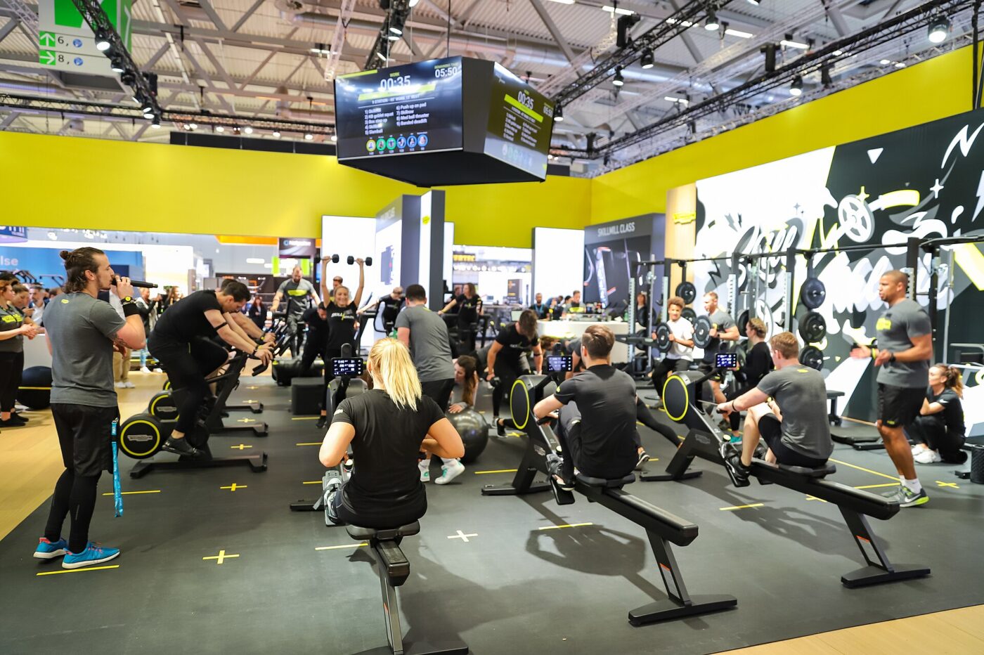 People row on indoor rowing machines during a coached group workout in a large exhibition hall