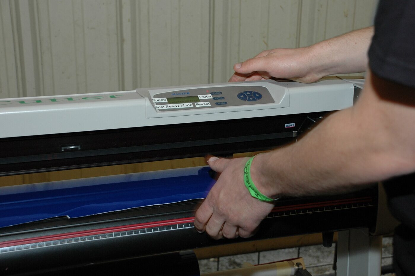 Hands feed a sheet of blue vinyl into a wide-format vinyl cutting machine