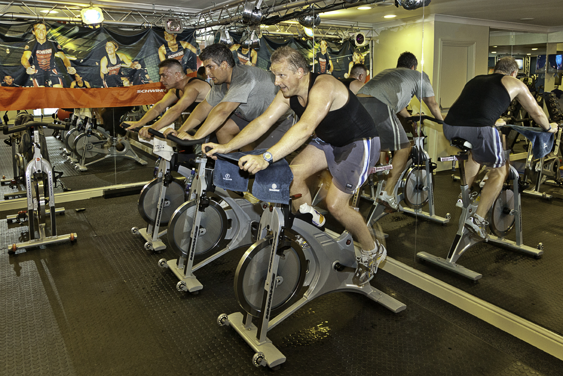 Riders pedal stationary spin bikes side by side during an indoor cycling class in a gym
