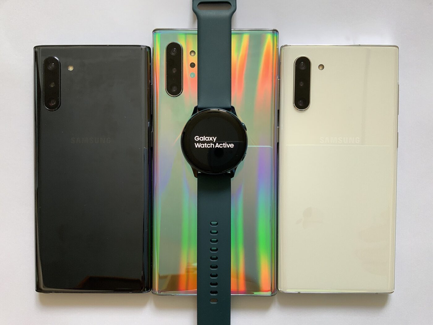 Round smartwatch with a dark green band laid across three smartphones on a white surface