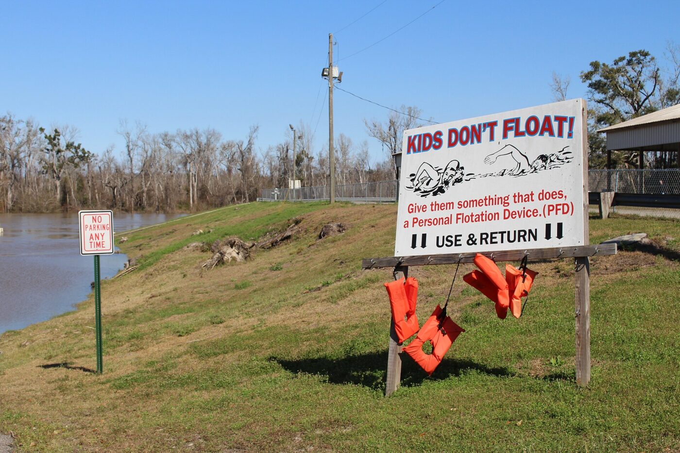 Orange life jackets hang from a public loaner sign beside a river on a sunny day.
