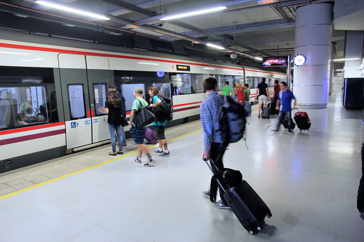 Travelers with rolling suitcases and backpacks walk along a train station platform