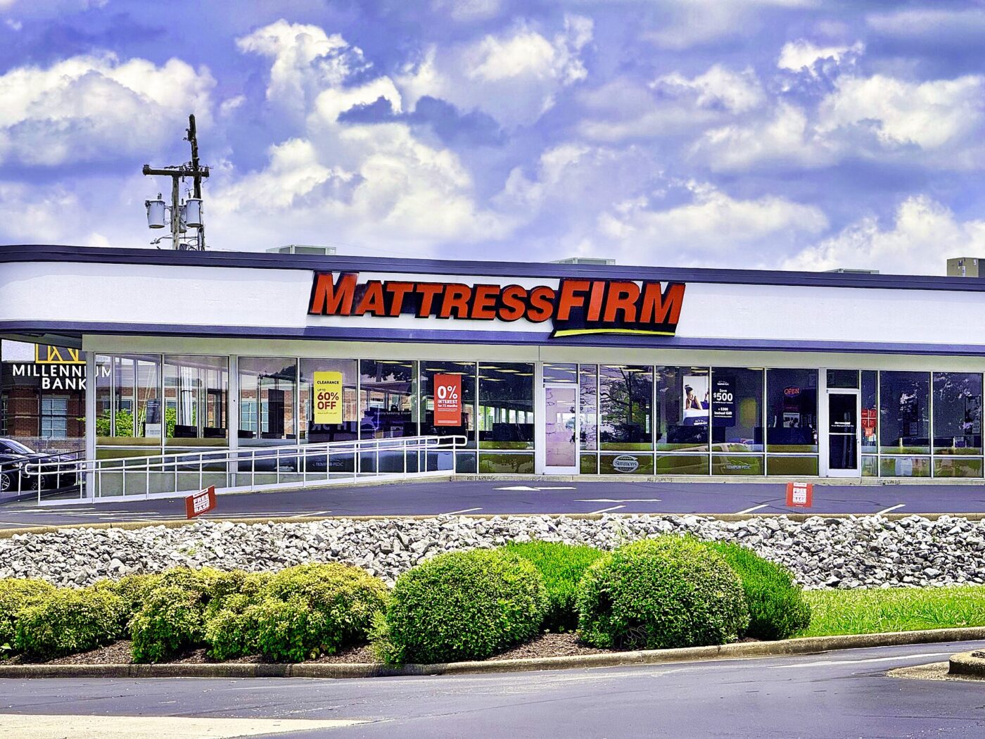A Mattress Firm store with sale signs in its windows, set behind a landscaped parking lot