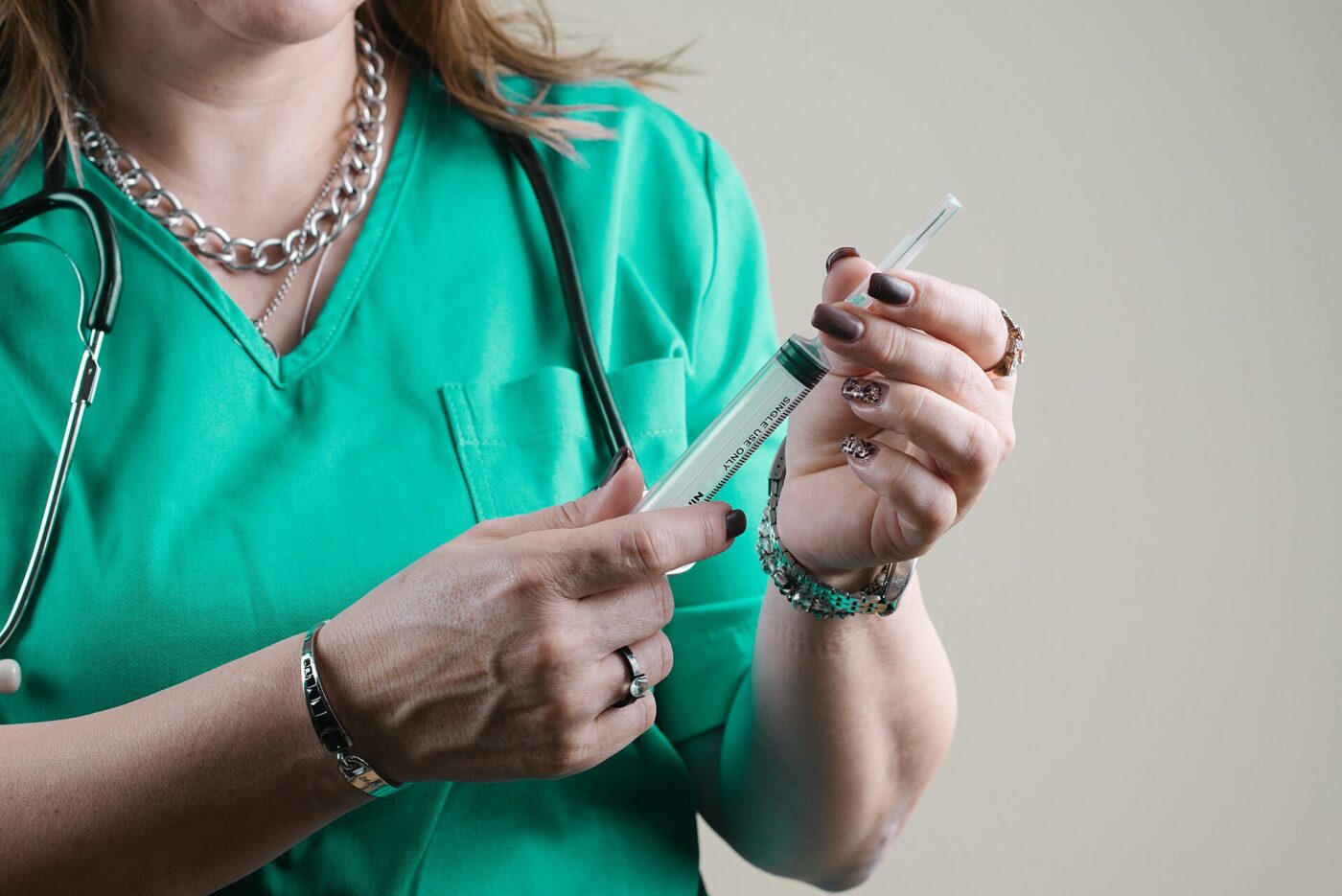 A healthcare worker in a green scrub top with a stethoscope holds a syringe