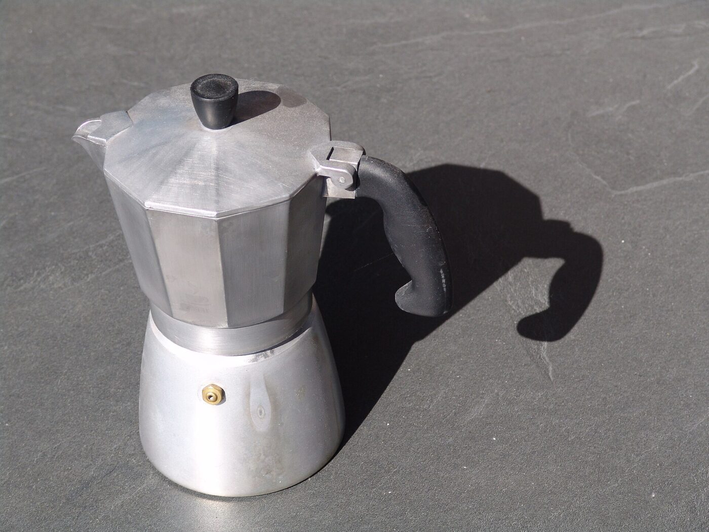 An aluminum stovetop moka pot on a gray stone surface, casting a long shadow.