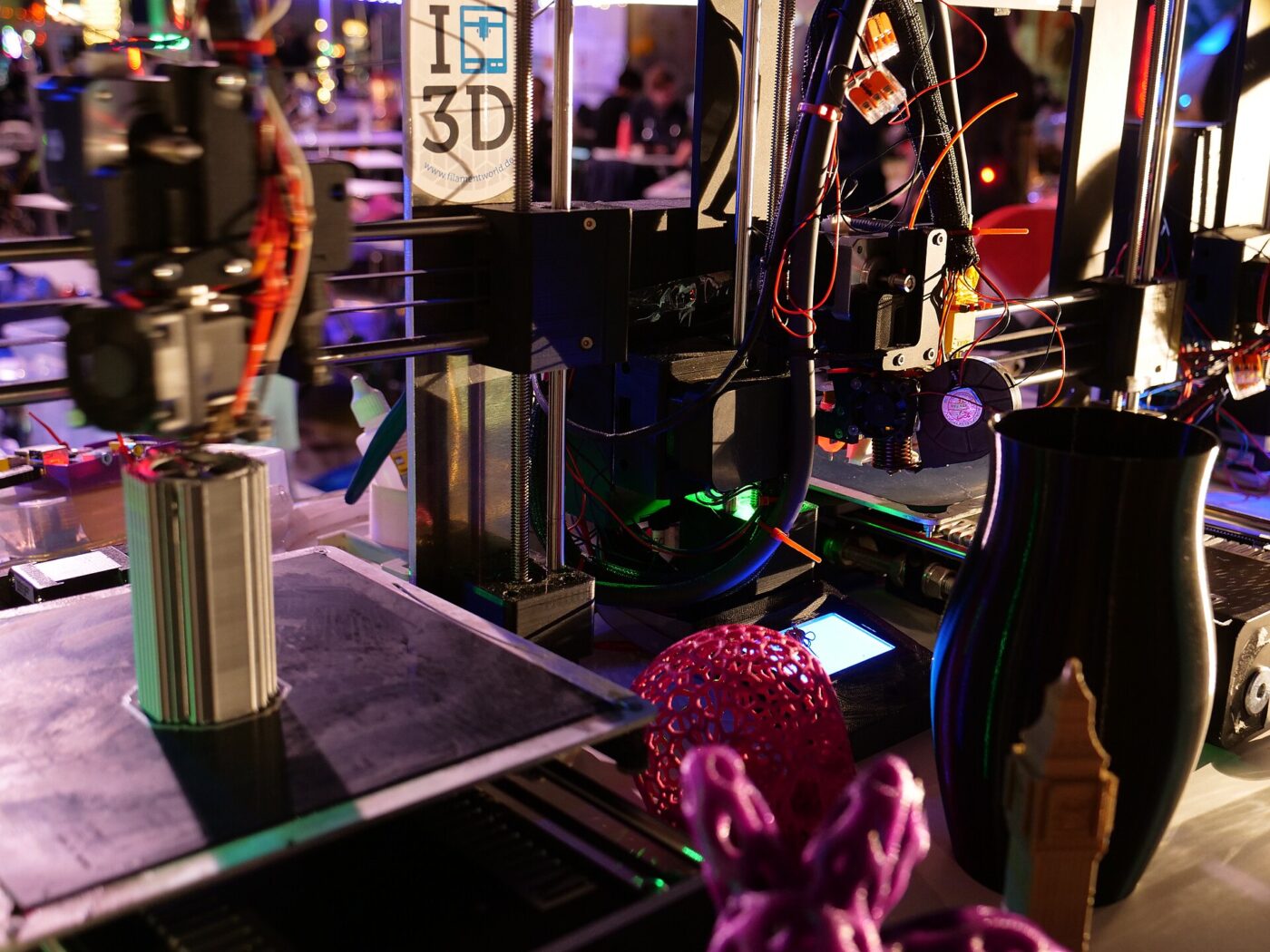 Open-frame 3D printers on a table beside finished prints in pink, black and tan plastic