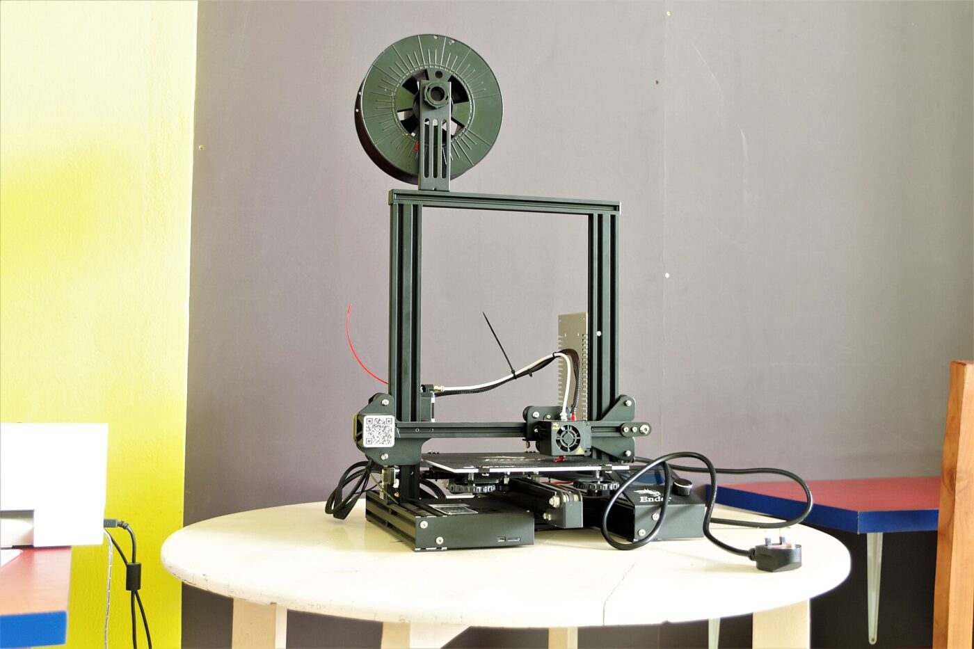 An open-frame desktop 3D printer with a filament spool mounted on top, set on a round white table