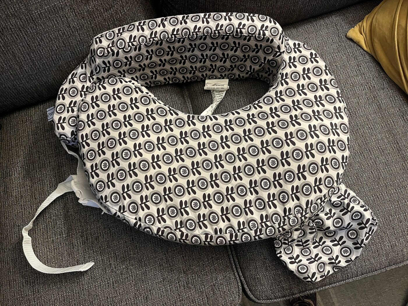 A wraparound nursing pillow with a fabric strap and buckle lying on a sofa.