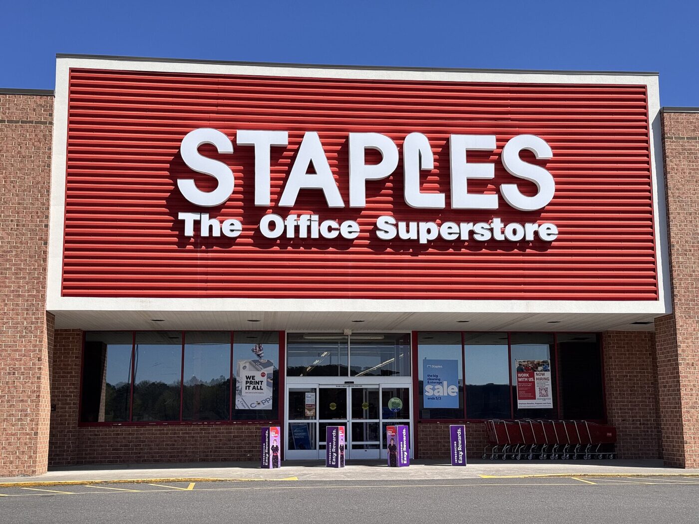 The storefront of a Staples office superstore, its red sign above glass entrance doors