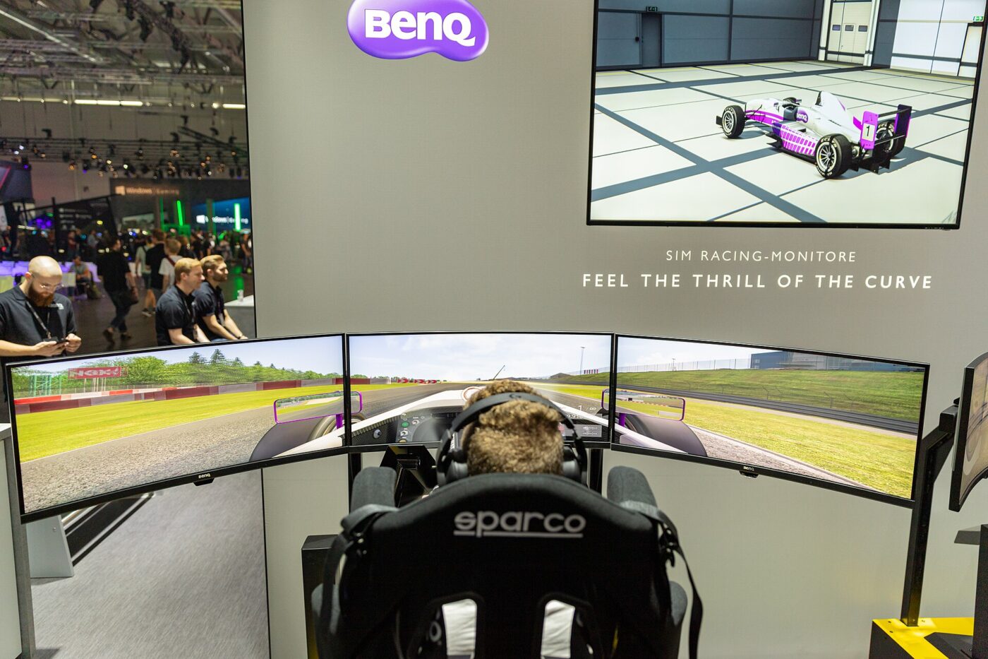 A player sits in a racing simulator seat facing three curved gaming monitors at a trade show booth
