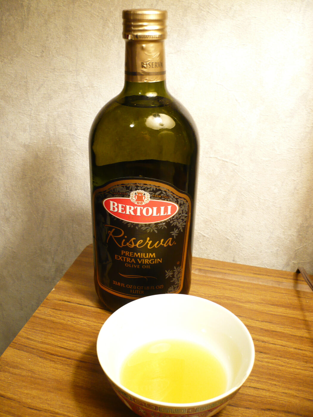 A dark glass extra virgin olive oil bottle with its front label, beside a bowl of poured oil